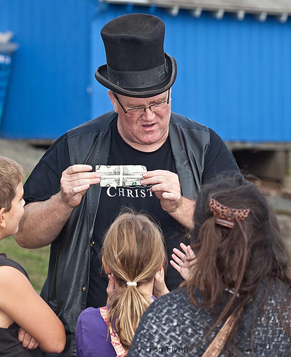 A magician wearing a black top hat performs a trick with a dollar bill for a small audience.